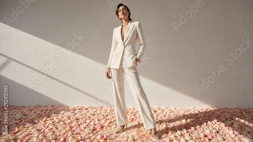 Elegant fashion portrait of a woman in a white tailored suit standing on a carpet of pink petals. Ideal for editorials, luxury campaigns, bridal branding and modern advertising.