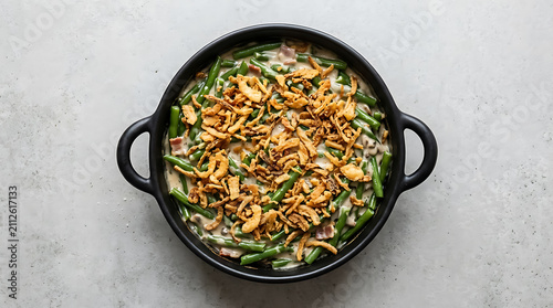 View of a creamy green bean casserole in a black baking dish topped with crispy fried onions on a light gray textured concrete background