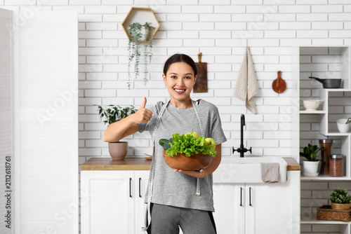Young woman in sportswear, with measuring tape and healthy food in kitchen. W...