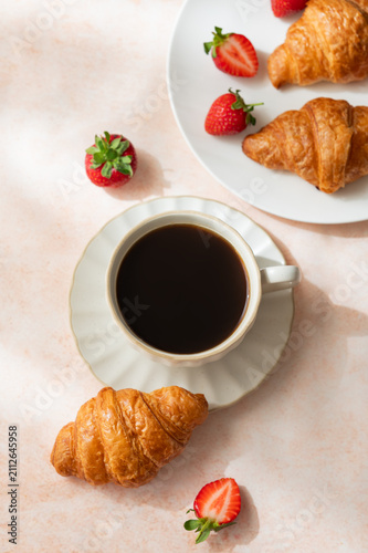 Baked croissants, cup of espresso coffee and strawberries on a bright textured background.