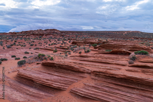 Sculptured red sandstone desert landscape at Horseshoe Bend near Page, Arizona. Layered rock formations and sparse vegetation. Mountains and cloudy sky in the background. 
