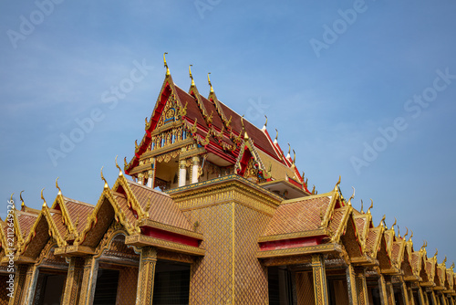 Ornate, multi-tiered red and gold roof of the Buddhist temple Wat Panthai Norasing in Bangkok, Thailand. Wispy clouds and blue sky behind. 
