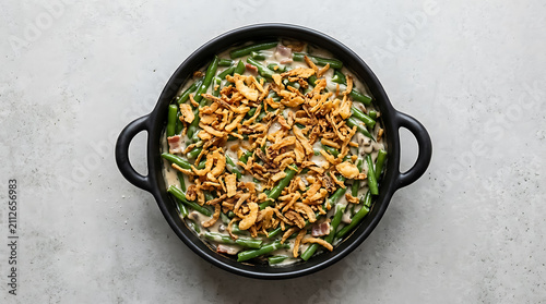 View of a creamy green bean casserole in a black baking dish topped with crispy fried onions on a light gray textured concrete background