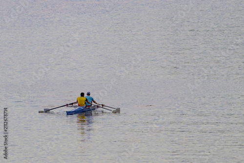 Two people row a small boat on calm water. They use oars to propel the vessel forward. The water is gentle with subtle ripples. The rowers wear bright tops for visibility. Their motion suggests steady