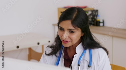 Young indian female doctor providing a remote consultation via video call from her office, explaining treatment and giving medical advice to a patient with a friendly and reassuring smile