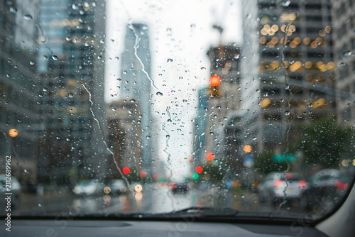 Raindrops on a car windshield with a blurred city street view in the background, showing urban buildings and traffic lights during a rainy day