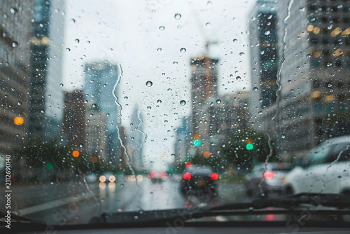 Raindrops on a car windshield with a blurred city street view in the background, showing urban buildings and traffic lights during a rainy day