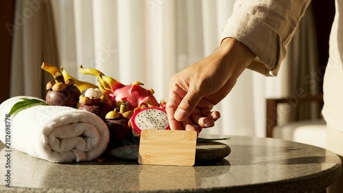 Hand Placing Wooden Sign Next To A Vibrant Tropical Fruit Arrangement On Stone Surface In A Wellness Hospitality Setting