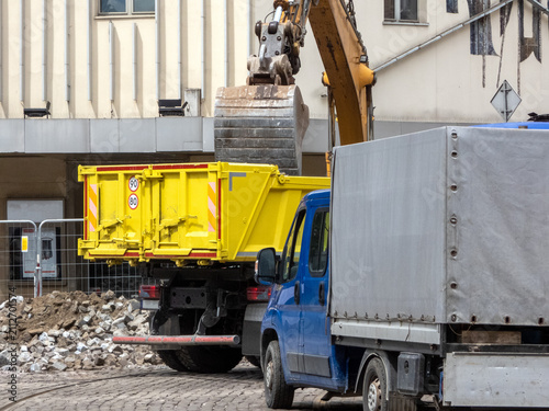 A yellow hydraulic excavator next to a large, yellow RORO (Roll-On/Roll-Off) skip container. commercial waste removal services. A blue truck  for hauling away the debris. Demolition operations. 