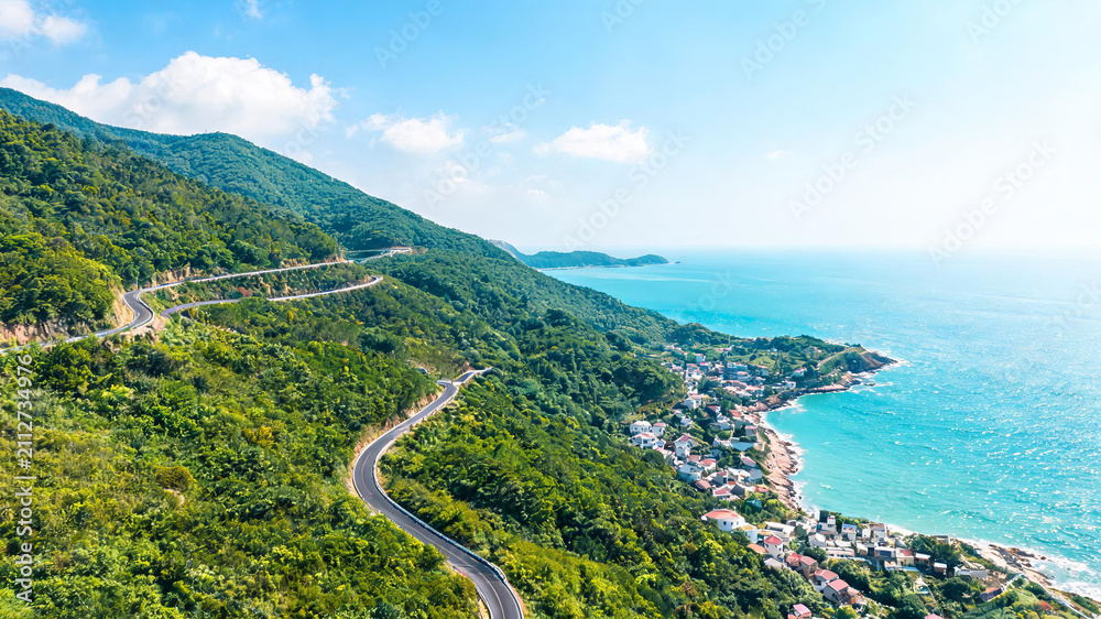 Fototapeta premium A scenic winding road cuts through green mountains beside a turquoise sea with a small coastal village under clear blue sky.