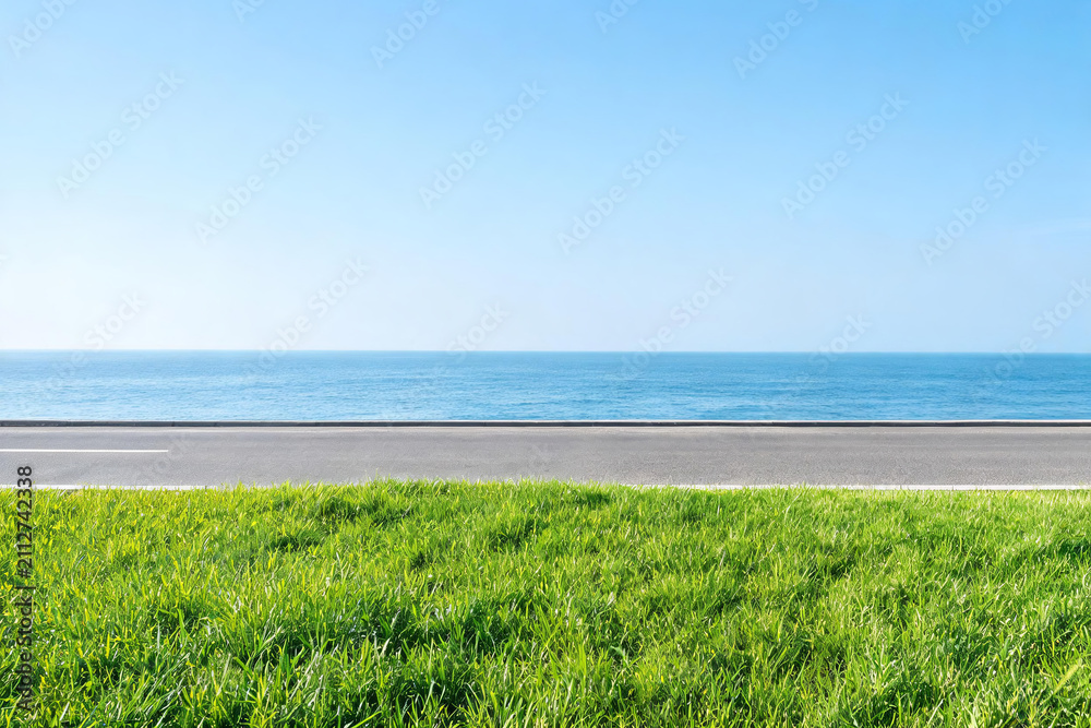 Obraz premium A straight asphalt road runs between lush green grass and a calm blue sea under a clear bright sky.