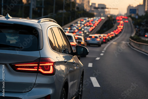A line of cars stuck in heavy traffic on an urban highway during sunset, with glowing red taillights stretching into the distance.