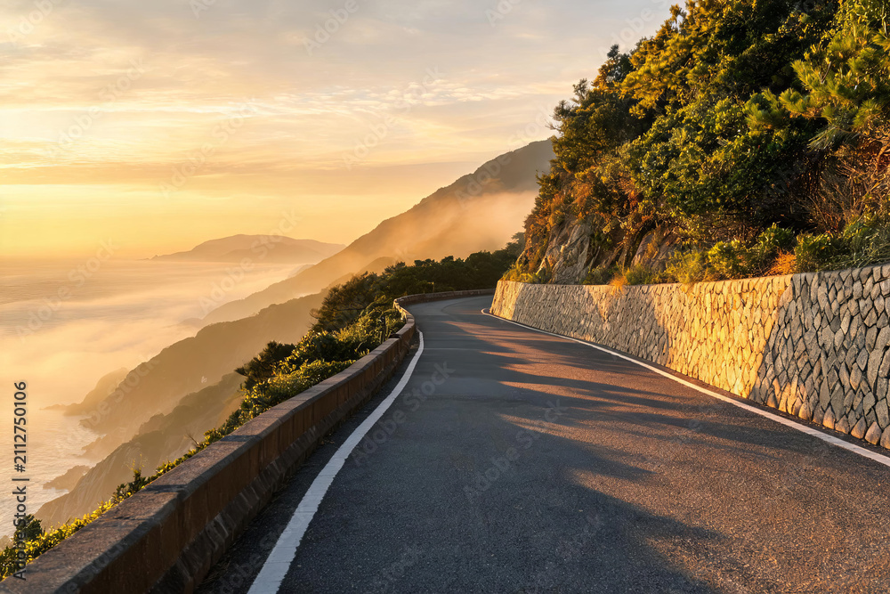 Obraz premium A winding mountain road cuts through coastal terrain at sunrise, with mist drifting over distant peaks and warm golden light illuminating the landscape.