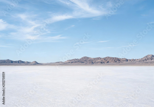 A wide, flat saltpan stretches toward distant rocky mountains under a bright, cloud-dotted blue sky, showing an arid natural landscape.