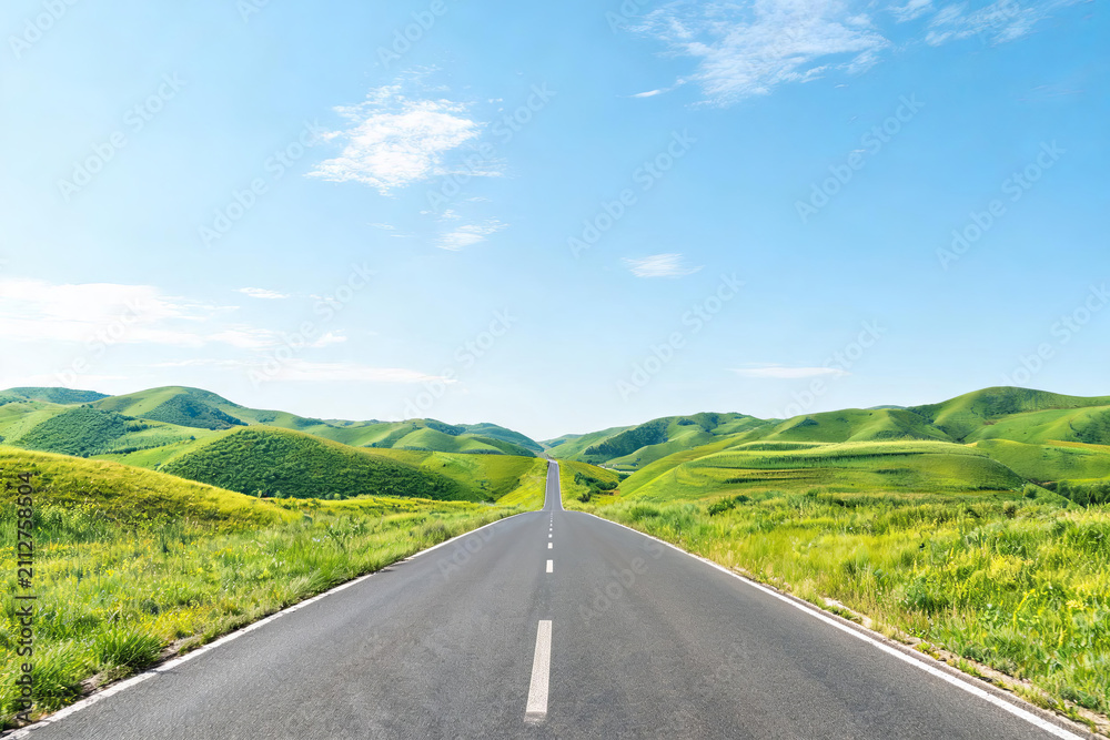 Obraz premium A straight asphalt highway stretches through lush green rolling hills under a clear bright blue sky with scattered clouds.