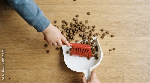 A young toddler sweeping spilled pet food into a dustpan with a red brush on a wooden floor