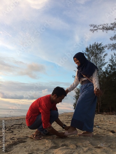 A man in a red shirt is putting on sandals for his girlfriend on a beach.