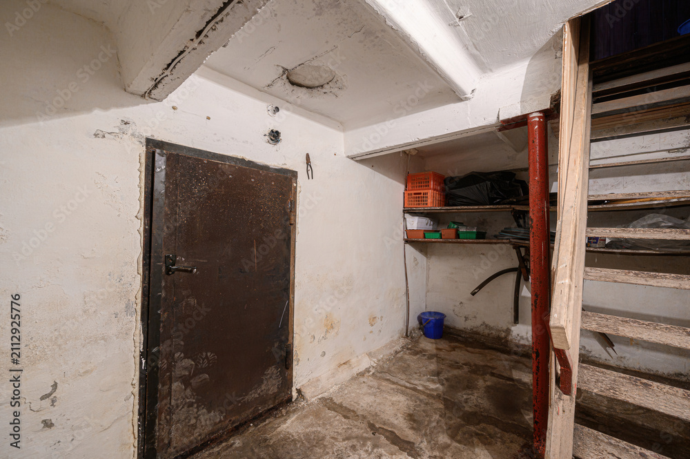Fototapeta premium cramped utility corner features a dark metal door, wall‑mounted shelves with plastic crates and containers, a blue bucket on the stained concrete floor, and a wooden staircase
