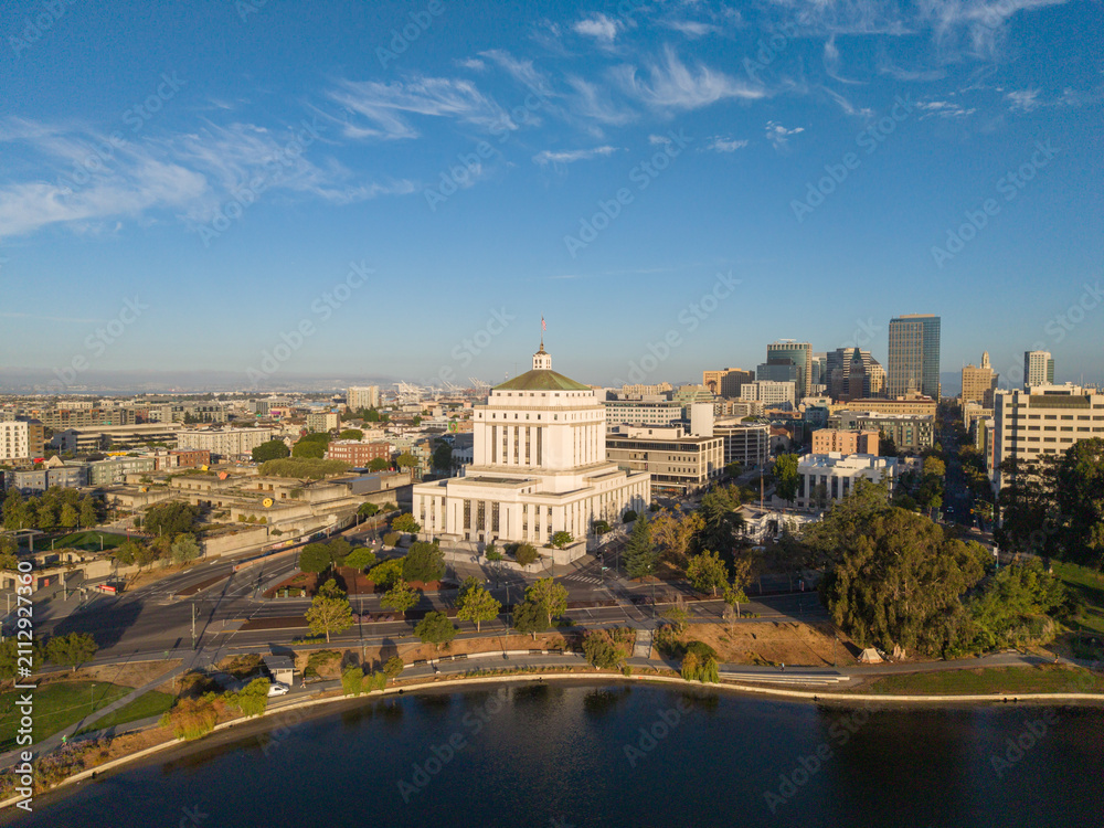 Fototapeta premium Aerial view displaying the iconic Oakland City Hall building next to Lake Merritt with the downtown skyline under a clear blue sky, highlighting urban landscape and government architecture