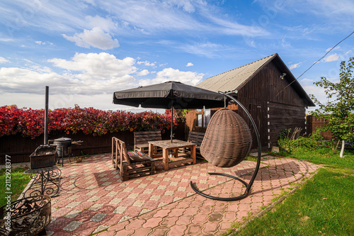 cozy backyard patio features wooden table and benches, black umbrella, wicker egg chair on metal stand, fire pit, rustic wooden house amid red‑leaf hedges and green lawn