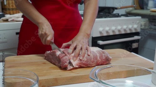 A person is cutting raw meat on a wooden cutting board in a kitchen setting, with a stove and countertop visible in the background. The scene is captured from a close-up, slightly elevated.