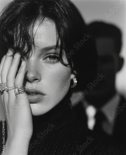 Female model with dark hair and freckles poses with hand on face, wearing rings, while a blurred figure in a suit is visible in the background