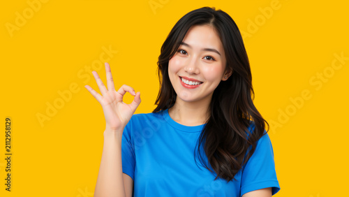 Happy young Asian woman showing OK hand gesture. Smiling female model in blue t-shirt on vibrant yellow background. Approval and satisfaction concept