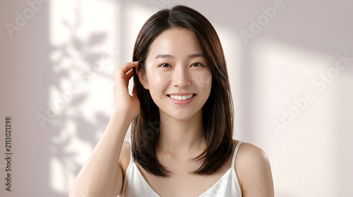 Bright smiling young woman with smooth dark hair wearing a casual white tank top in a minimalistic indoor setting with natural