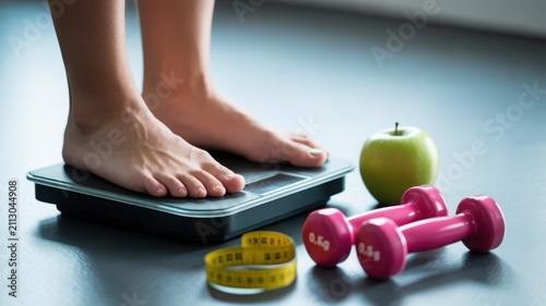 Person's bare feet on a digital weighing scale with dumbbells, apple, and measuring tape, representing weight loss and healthy lifestyle