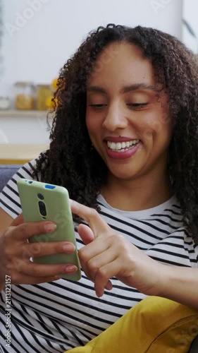 Vertical HD video of young African American woman relaxing on sofa while using smartphone at home. Smiling young woman laughing while browsing social media content in a modern apartment