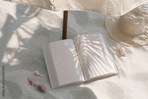 Blank white book mockup in sand under beach umbrella with straw hat