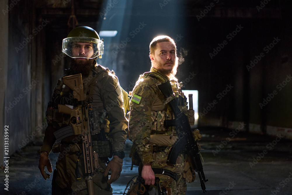Obraz premium Two Soldiers in Tactical Gear Standing Back-to-Back Inside Dimly Lit Industrial Building