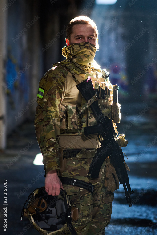 Obraz premium Masked Soldier In Camouflage Tactical Gear Holding Rifle And Helmet In Urban Setting