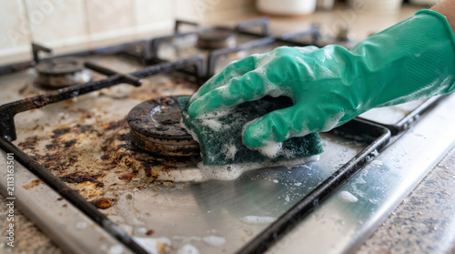 Person wearing rubber gloves scrubbing greasy dirty gas stove top with sponge and soap foam