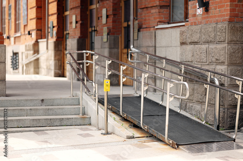 Accessible building entrance with non-slip wheelchair ramp, stainless steel handrails, and yellow assistance call button.