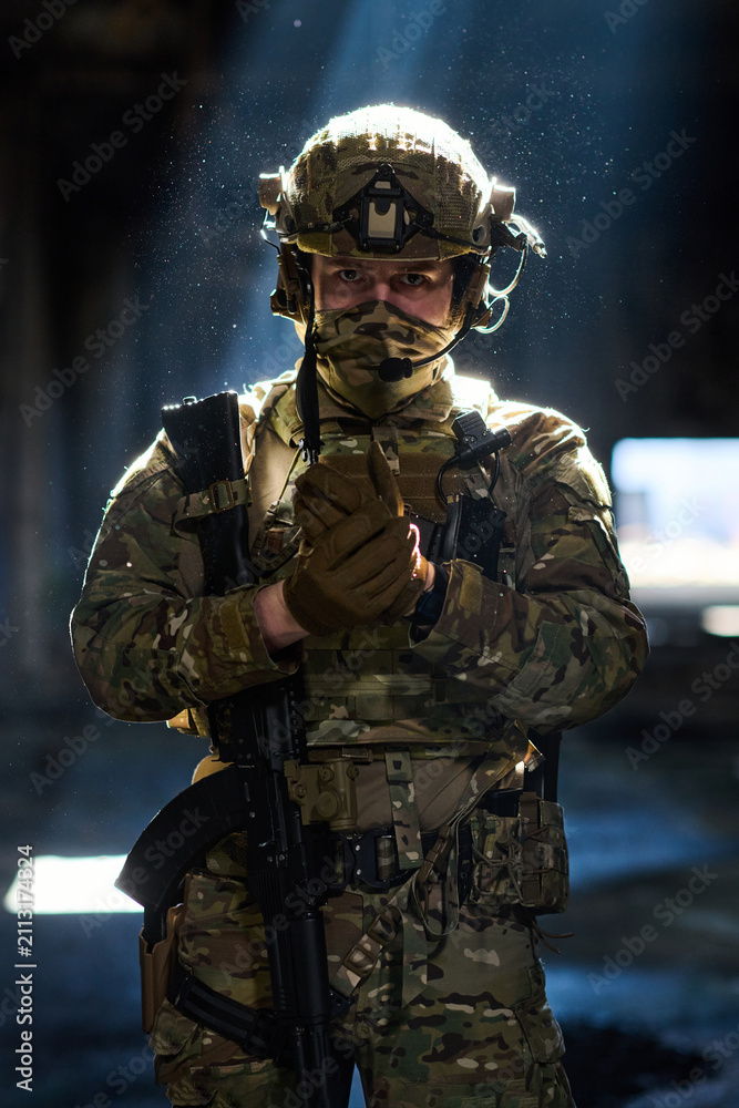Obraz premium Armed Soldier in Camouflage Tactical Gear With Rifle and Helmet Standing in Dim Light
