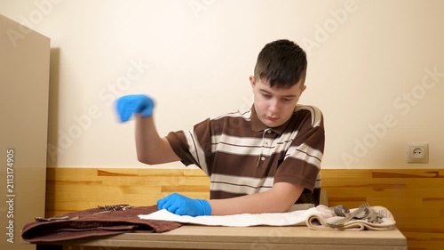 Teenager wearing protective gloves is polishing a metal spoon with a towel while carefully cleaning cutlery for table setting.