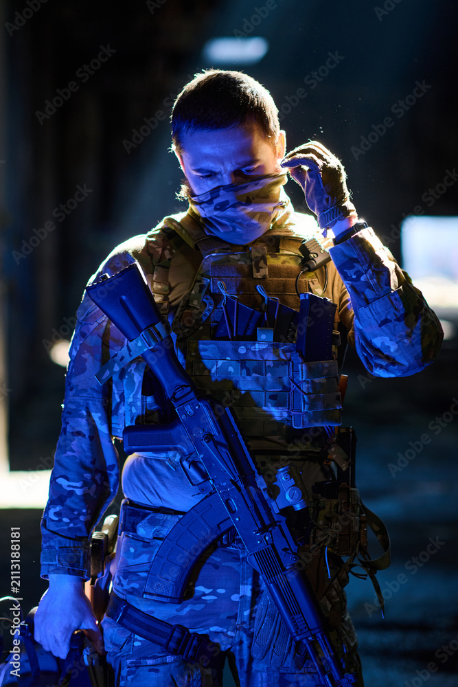 Obraz premium Armed Soldier in Tactical Camouflage Gear Adjusting Face Mask While Holding Assault Rifle in Low Light
