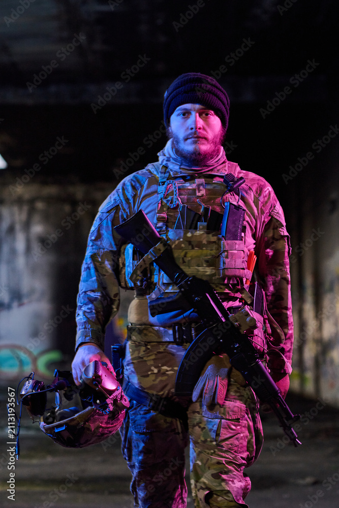 Obraz premium Armed Soldier In Camouflage Tactical Gear Holding Helmet And Assault Rifle In Urban Setting