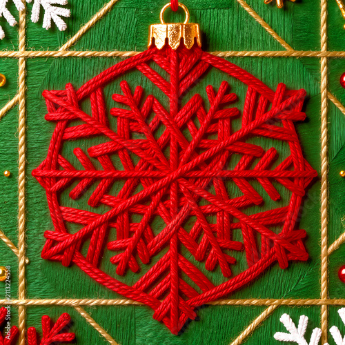 A vibrant square tile illustration featuring a handcrafted red hexagonal snowflake ornament set against a green woven background with golden grid lines.