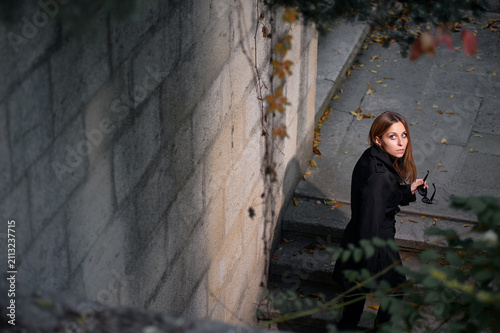 A lone woman in a black coat stands on outdoor stone steps beside a concrete wall, looking up with a contemplative, slightly startled expression amid fallen autumn leaves and urban shadows.