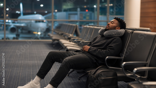 African american man sleeping on seat in airport lounge during flight delay. Traveler resting with neck pillow while waiting for departure. Long overnight layover or trip cancellation.