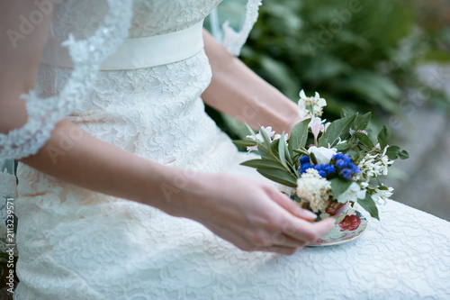 Bride wearing lace wedding dress delicately holds a small floral arrangement presented in a vintage teacup, evoking romantic, intimate, and vintage wedding styling with soft natural light and detail.
