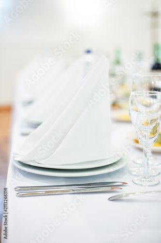 Elegant white place settings featuring folded napkins, stacked plates, silver cutlery and sparkling glassware on a linen-covered table for a wedding reception, formal banquet or upscale catered event.