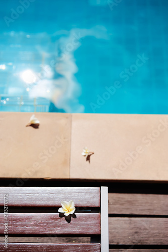 A single plumeria rests on a wooden pool lounge beside bright blue water and tiled edge, capturing warm summer light, quiet relaxation and a peaceful tropical holiday mood.
