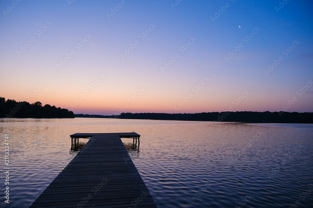 Fototapeta premium Moscow. Russia. 25.07.2026 Wooden pier extending into a calm lake at dusk. Serene sunset landscape with moonlight and twilight sky over water. Peaceful nature scene for travel and relaxation concept b