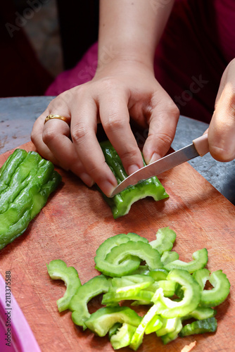 Close-up of woman's hands slicing fresh bitter melon on a wooden cutting board while preparing a healthy homemade meal. Home cooking, vegetable preparation, and nutritious food. Vertical
