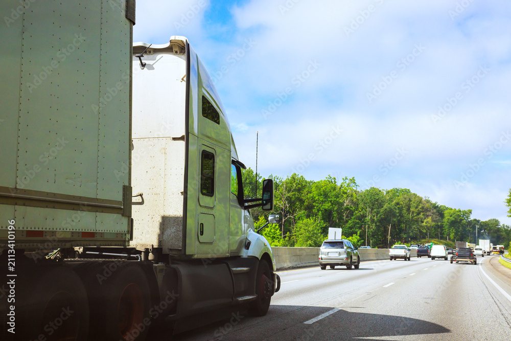 Obraz premium Large truck drives on busy highway with other cars during day trees along roadside.
