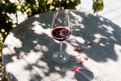 A glass of red wine sits on a textured surface with scattered flower petals and dappled sunlight