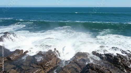 Ocean surf breaks over jagged coastal rocks while deep blue waves roll toward the horizon under a clear sky.